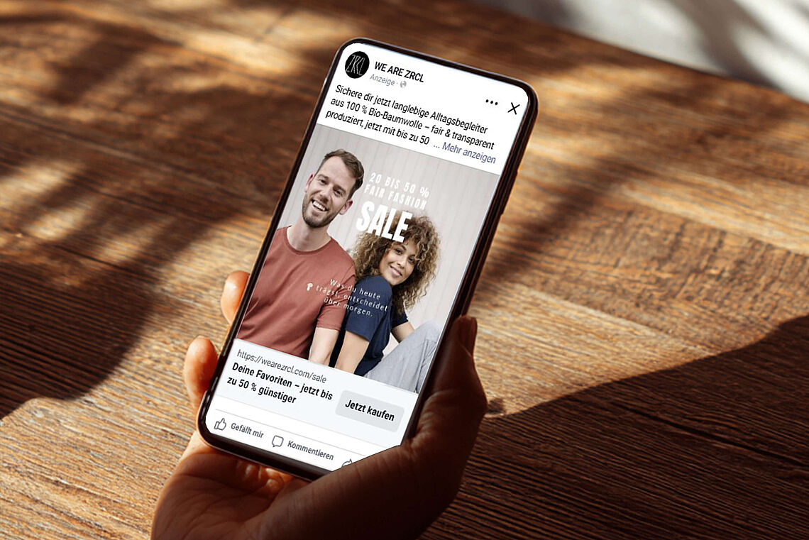 Eine Hand hält ein Smartphone, auf dem eine Facebook-Werbeanzeige von WE ARE ZRCL zu sehen ist. Zwei Personen tragen T-Shirts, daneben steht der Hinweis auf einen Sale mit bis zu 50 % Rabatt.
