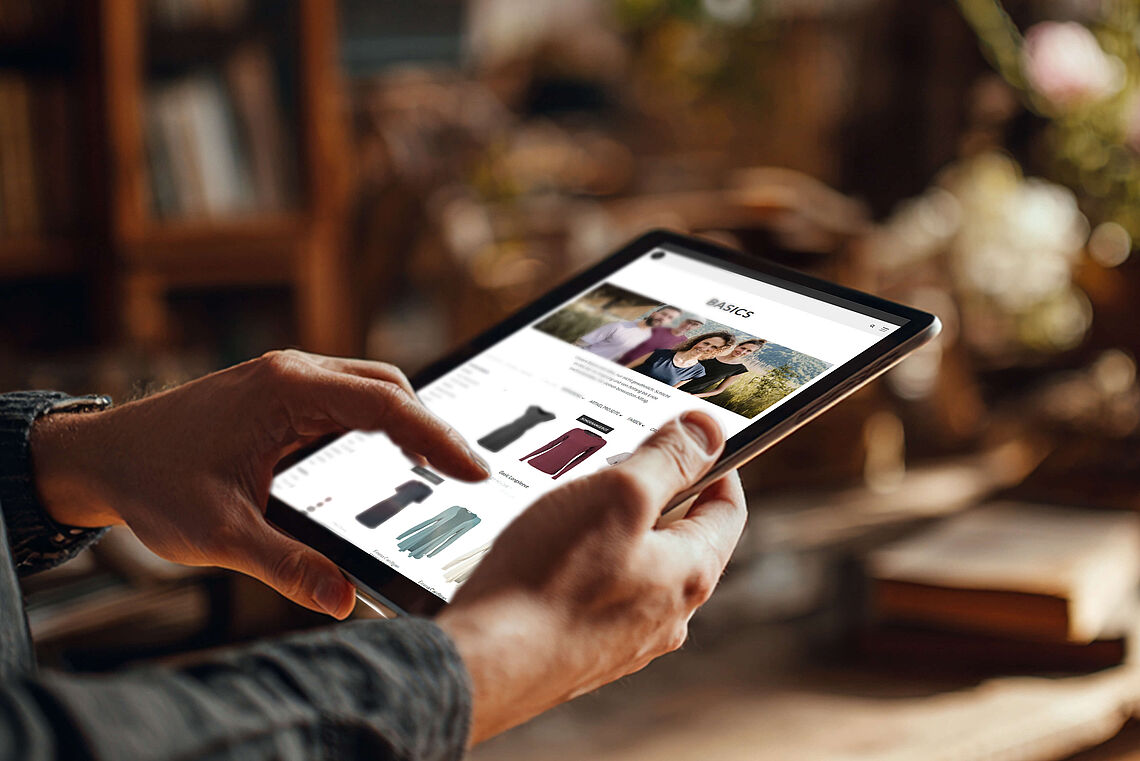 Eine Person hält ein Tablet, auf dem die Website von WE ARE ZRCL geöffnet ist. Die Seite zeigt eine Produktübersicht der Kategorie ‚Basics‘ mit mehreren Kleidungsstücken.